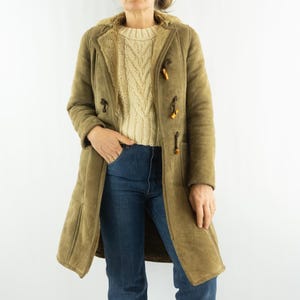 May include: An olive-green long coat with a shearling lining and wooden toggle fastenings. The coat is worn over a cream cable-knit sweater and blue jeans. The coat's length extends to mid-calf, offering warmth and style.
