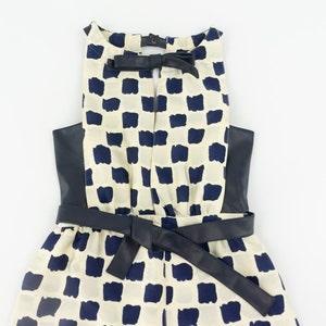 May include: A white and navy blue checkered dress with a black leather belt and a black leather trim around the neckline. The dress has a bow at the back of the neckline.