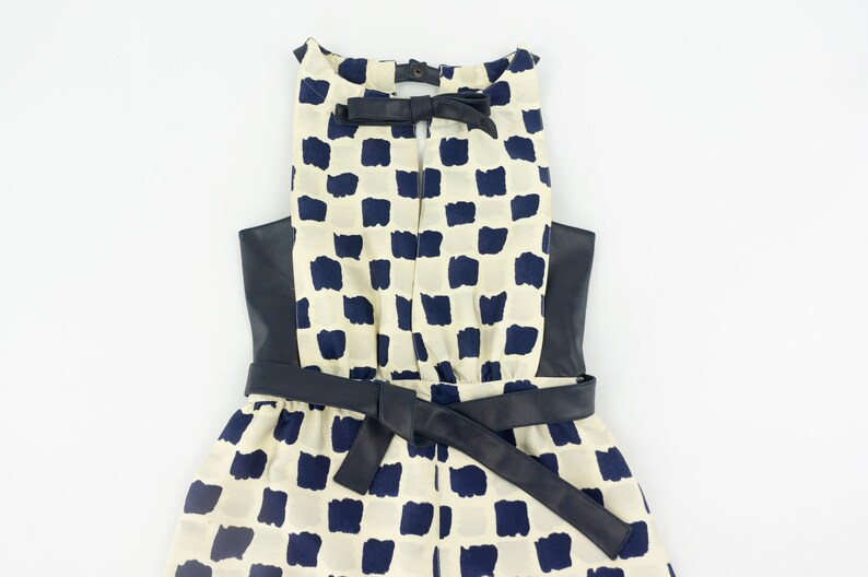 May include: A white and navy blue checkered dress with a black leather belt and a black leather trim around the neckline. The dress has a bow at the back of the neckline.
