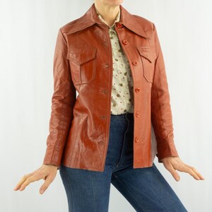 May include: A vintage, reddish-brown leather jacket with a collared neckline and button-down front. The jacket features two chest pockets and is paired with a patterned blouse and blue jeans. A classic outerwear piece.