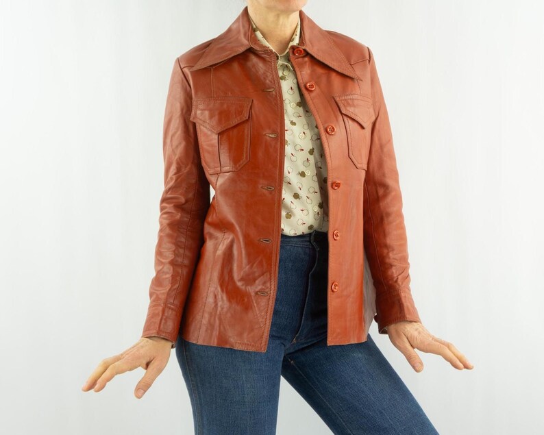 May include: A vintage, reddish-brown leather jacket with a collared neckline and button-down front. The jacket features two chest pockets and is paired with a patterned blouse and blue jeans. A classic outerwear piece.