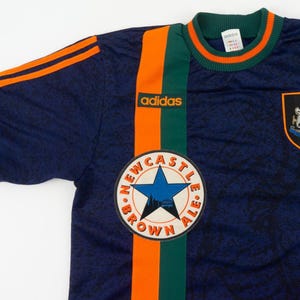 May include: Navy blue Adidas soccer jersey with orange and green stripes. Features the Newcastle Brown Ale logo and a team crest. The jersey has orange stripes on the sleeves and the Adidas logo.