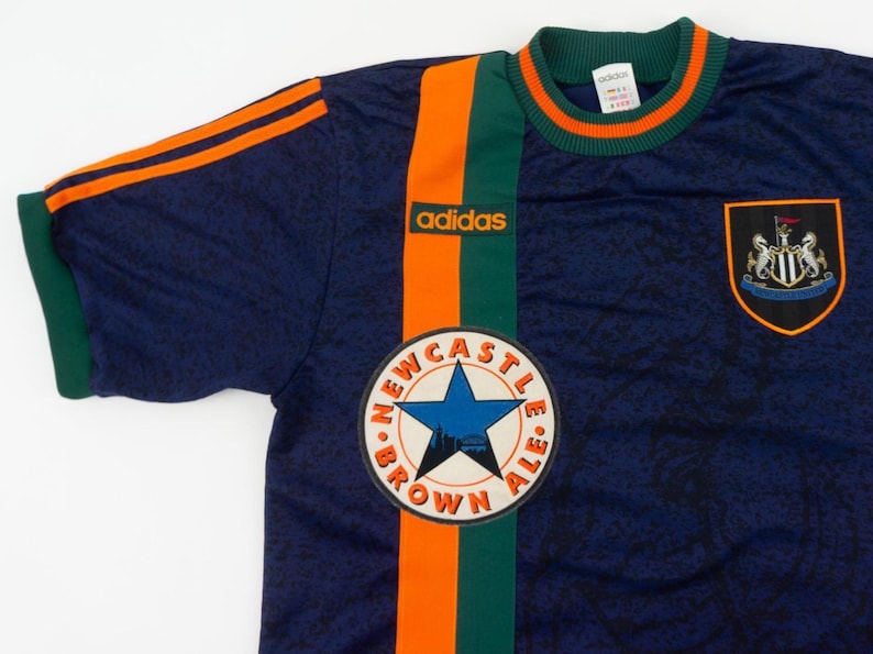 May include: Navy blue Adidas soccer jersey with orange and green stripes. Features the Newcastle Brown Ale logo and a team crest. The jersey has orange stripes on the sleeves and the Adidas logo.