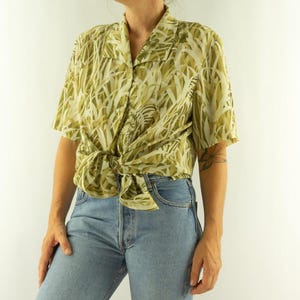 Pode incluir: Uma camisa de manga curta com botões com um padrão verde e creme, amarrada na cintura. A camisa é combinada com jeans azul claro. O design da camisa assemelha-se a fios de erva.