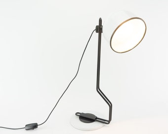 Könnte beinhalten: Eine weiß-schwarze Schreibtischlampe mit rundem Lampenschirm und schwarzem Sockel. Der Lampenschirm hat eine goldfarbene Einfassung. Die Lampe ist über ein schwarzes Kabel mit einer Steckdose verbunden und hat einen Ein-/Ausschalter.