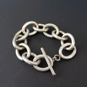 Peut inclure: Bracelet en argent avec des maillons ovales. Le bracelet est doté d'une fermeture à bascule. Le bijou est présenté sur un fond sombre. Un accessoire élégant pour un usage quotidien.