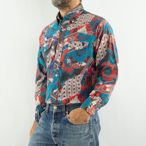Peut inclure: Une chemise à manches longues à motifs rouges, bleus et noirs. Elle est ornée de deux poches de poitrine et d'un col classique. La chemise est portée avec un jean bleu. Le motif est composé de formes variées.
