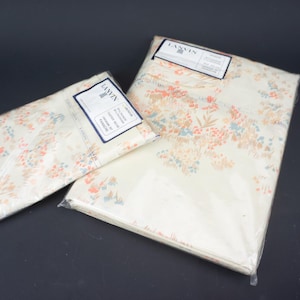 Puede incluir: Dos juegos de tela estampada con motivos florales, empaquetados en plástico transparente. La tela presenta un fondo crema con un delicado estampado de flores melocotón, azules y verdes. Cada paquete tiene una etiqueta con la marca "LANVIN".