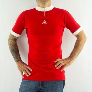 May include: A bright red short-sleeve shirt featuring a white mock neck collar with a small front zipper. The shirt has white trim on the sleeve cuffs and a small white triangular logo with a rooster graphic centered on the upper chest area.