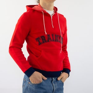 K&ouml;nnte beinhalten: Roter Kapuzenpullover mit einem dunkelblauen "TRAINING"-Logo auf der Vorderseite. Der Pullover hat einen halben Rei&szlig;verschluss und einen dunkelblauen Saum am unteren Rand.