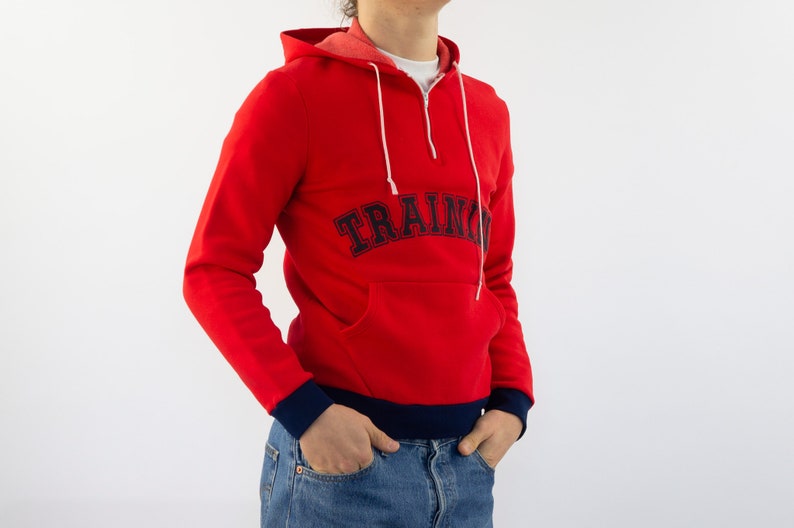 K&ouml;nnte beinhalten: Roter Kapuzenpullover mit einem dunkelblauen "TRAINING"-Logo auf der Vorderseite. Der Pullover hat einen halben Rei&szlig;verschluss und einen dunkelblauen Saum am unteren Rand.