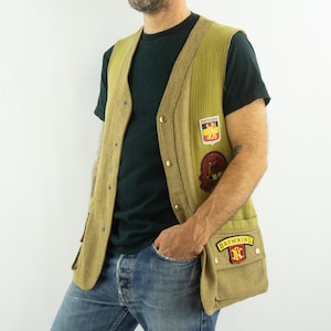 May include: A vintage olive green vest with a tan trim and pockets. The vest features several patches, including a Browning logo. The vest is buttoned up and has a classic, outdoorsy style.