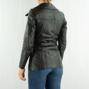 May include: A black leather jacket with a belted waist and a notched collar. The jacket has long sleeves and a tailored fit. The model is wearing blue jeans. The jacket is a classic outerwear piece.