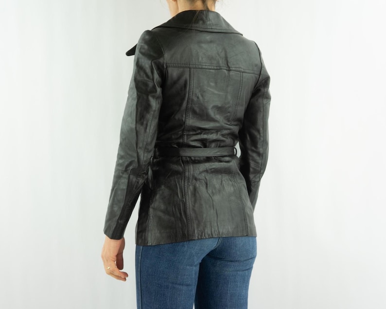May include: A black leather jacket with a belted waist and a notched collar. The jacket has long sleeves and a tailored fit. The model is wearing blue jeans. The jacket is a classic outerwear piece.