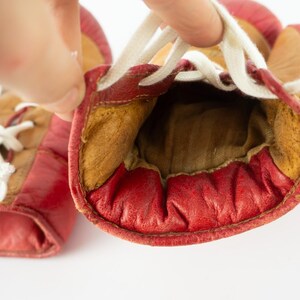 May include: A pair of vintage red leather boxing gloves with white laces. The gloves have a worn appearance, with visible stitching and a tan interior. The gloves are held open, showcasing the inside padding.