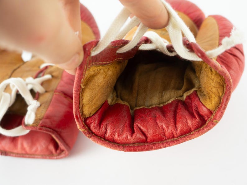 May include: A pair of vintage red leather boxing gloves with white laces. The gloves have a worn appearance, with visible stitching and a tan interior. The gloves are held open, showcasing the inside padding.