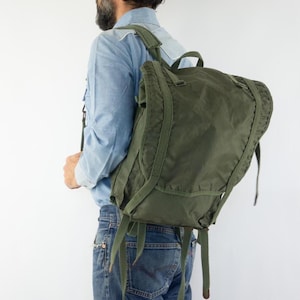 Könnte beinhalten: Olivgrüner Rucksack mit mehreren Riemen und einem Klappenverschluss. Der Rucksack wird über der Schulter getragen. Die Tasche ist aus strapazierfähigem Material gefertigt und scheint von Vintage- oder Militärstil inspiriert zu sein. Die Person trägt ein blaues Hemd und Jeans.