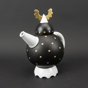 Peut inclure: Une théière en céramique noire et blanche avec des détails dorés. La théière a un corps rond avec des pois dorés, un bec et une anse blancs, et un élément décoratif doré sur le couvercle. La base est blanche.