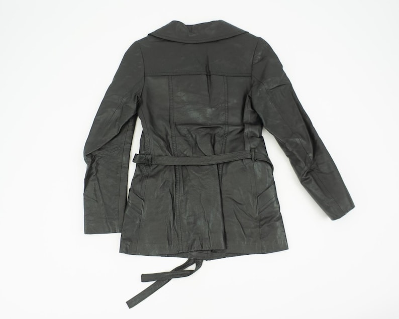 May include: A black leather jacket with a notched collar, long sleeves, and a belted waist. The jacket has a textured appearance and a tie closure at the bottom. The back of the jacket is visible against a white background.