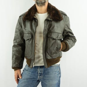 Peut inclure: Veste de bombardier en cuir vert olive vintage avec un col en fourrure marron. La veste a deux poches avant et des poignets et un ourlet côtelés marron. Le modèle porte un jean bleu et un t-shirt blanc.