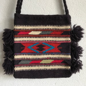 May include: A woven shoulder bag with a black background and a colourful geometric pattern. The pattern includes red, blue, white, and green shapes. The bag has a braided strap and black fringe on the sides.
