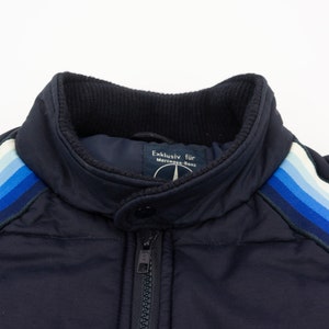May include: A navy blue jacket with a zipper closure and a white and blue striped detail on the sleeves. The jacket has a label that reads "Exclusiv fur Mercedes-Benz".