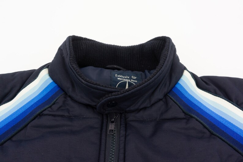 May include: A navy blue jacket with a zipper closure and a white and blue striped detail on the sleeves. The jacket has a label that reads "Exclusiv fur Mercedes-Benz".