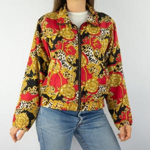 Op de afbeelding: Een rood, zwart en gouden gepatroneerd jack met een ritssluiting. Het jack heeft een luipaardprint met gouden accenten.