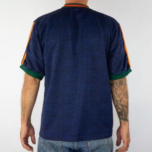 May include: A navy blue short-sleeve shirt with orange stripes on the sleeves and green trim. The shirt has a speckled pattern and a ribbed collar with orange and green stripes. The shirt is a vintage style.