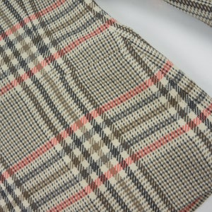Pu&ograve; includere: Primo piano di un tessuto con un classico motivo a quadri. Il design presenta una griglia di linee intersecanti nelle tonalit&agrave; del beige, marrone, nero e rosa. Il tessuto sembra far parte di un capo di abbigliamento, forse un cappotto o una giacca.