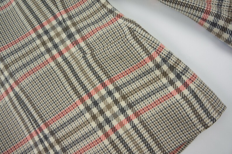 Pu&ograve; includere: Primo piano di un tessuto con un classico motivo a quadri. Il design presenta una griglia di linee intersecanti nelle tonalit&agrave; del beige, marrone, nero e rosa. Il tessuto sembra far parte di un capo di abbigliamento, forse un cappotto o una giacca.