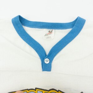 以下が含まれることがあります： Vネックと袖に青いトリムが付いた白いヴィンテージTシャツ。シャツは、ネックラインにボタン留めとタグが付いています。シャツはヴィンテージアイテムです。