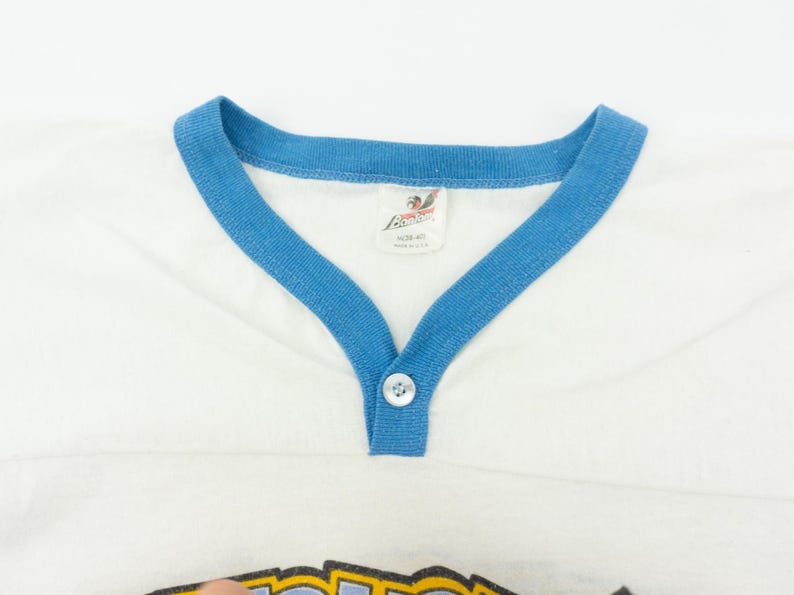以下が含まれることがあります： Vネックと袖に青いトリムが付いた白いヴィンテージTシャツ。シャツは、ネックラインにボタン留めとタグが付いています。シャツはヴィンテージアイテムです。