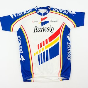 May include: A white, blue, and red Banesto cycling jersey. The jersey features the Banesto logo on the front and sleeves, with a design of red, yellow, and blue stripes. The jersey has a short zipper at the collar.