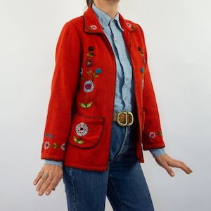 May include: A red embroidered jacket with floral designs. The jacket has a collar, long sleeves, and two pockets. The jacket is closed with a zipper. The jacket is worn over a light blue shirt and a brown belt.
