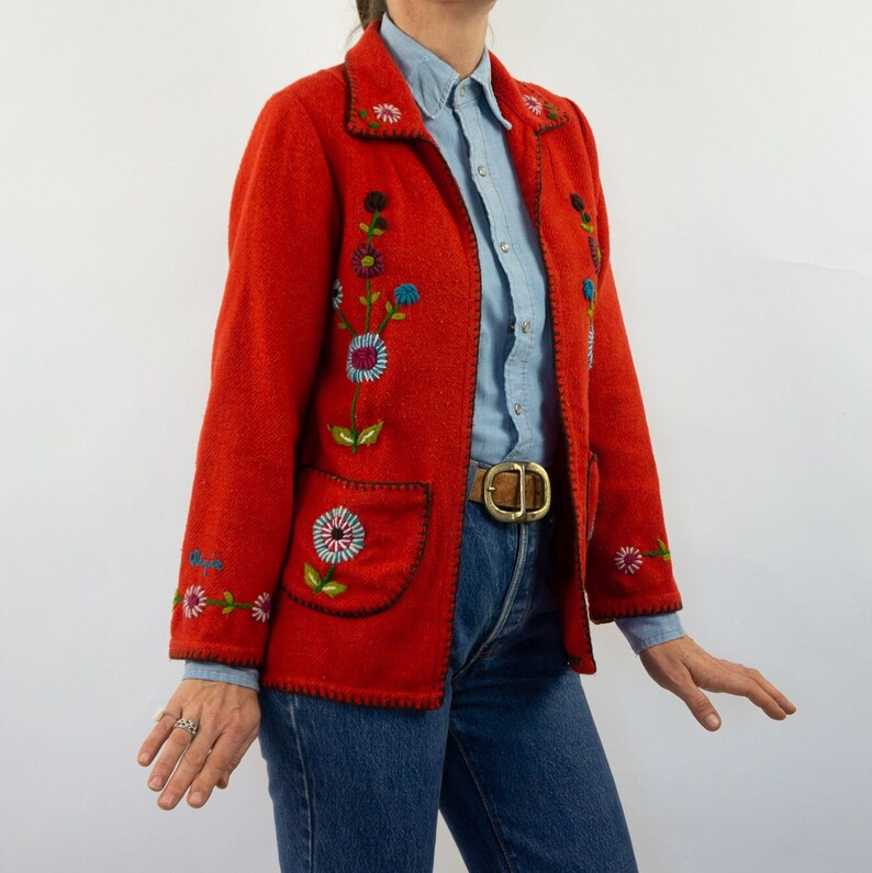 May include: A red embroidered jacket with floral designs. The jacket has a collar, long sleeves, and two pockets. The jacket is closed with a zipper. The jacket is worn over a light blue shirt and a brown belt.