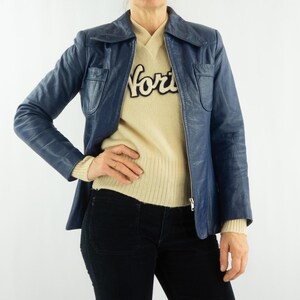 K&ouml;nnte beinhalten: Eine blaue Vintage-Lederjacke mit Kragen und Vordertaschen. Die Jacke wird &uuml;ber einem cremefarbenen Pullover mit dem Wort "for" in schwarzen Buchstaben getragen. Die Person tr&auml;gt eine schwarze Hose.
