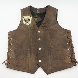 May include: Brown leather vest with a distressed finish, featuring a patch with text and a graphic. The vest has a button-up front, two front pockets, and lace-up sides. The vest is a classic biker style.