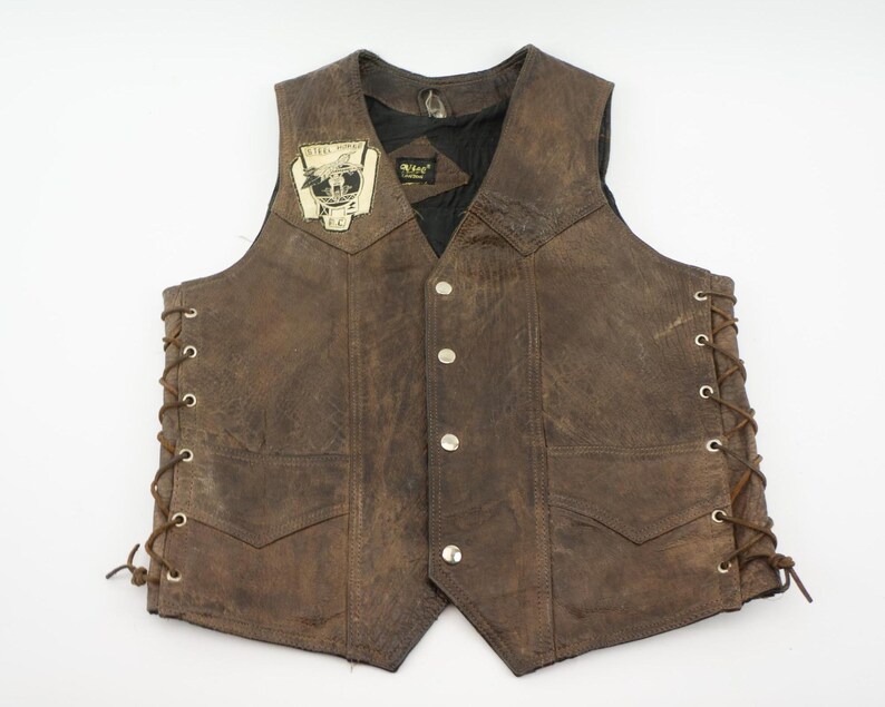 May include: Brown leather vest with a distressed finish, featuring a patch with text and a graphic. The vest has a button-up front, two front pockets, and lace-up sides. The vest is a classic biker style.