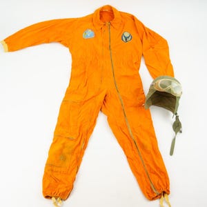 Peut inclure: Combinaison de vol orange vif avec fermeture éclair, écussons et poches. Comprend un casque d'aviateur vert avec des lunettes. La combinaison a un col et des poignets élastiques. Un ensemble d'aviation classique.