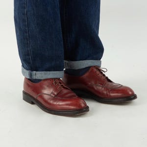 May include: A pair of brown leather lace-up shoes with a pointed toe. The shoes are worn with rolled-up blue jeans.