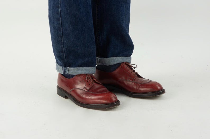 May include: A pair of brown leather lace-up shoes with a pointed toe. The shoes are worn with rolled-up blue jeans.