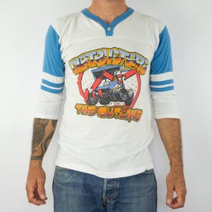 以下が含まれることがあります： レースカーのグラフィックと「Dirtbusters The Outlaws」の文字が入った白と青のラグランスタイルのシャツ。 シャツはVネックで、青いストライプの3/4袖です。 グラフィックは赤、黄、青です。