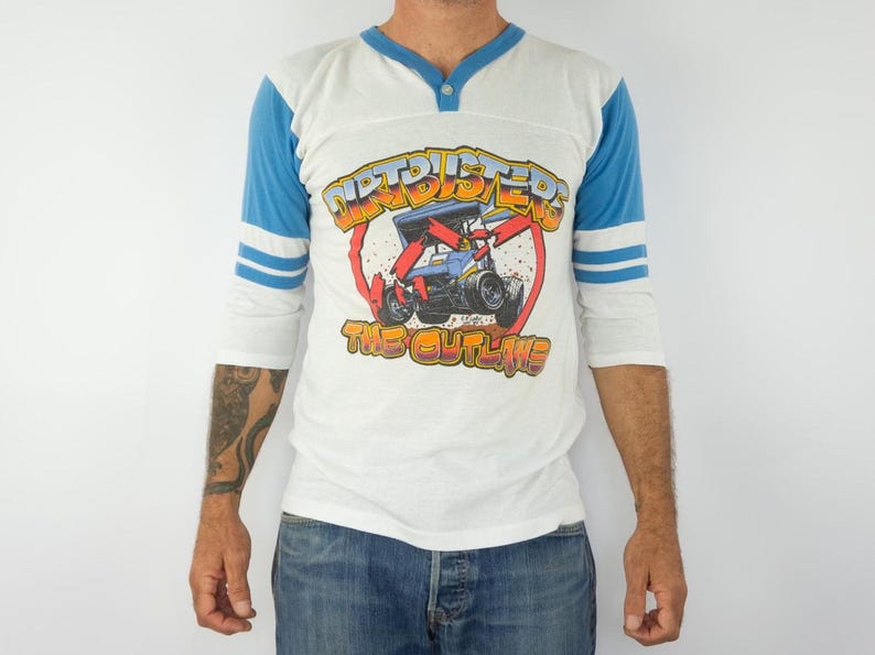以下が含まれることがあります： レースカーのグラフィックと「Dirtbusters The Outlaws」の文字が入った白と青のラグランスタイルのシャツ。 シャツはVネックで、青いストライプの3/4袖です。 グラフィックは赤、黄、青です。