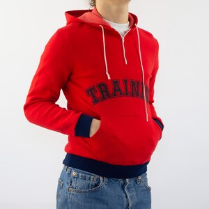 K&ouml;nnte beinhalten: Roter Kapuzenpullover mit dunkelblauem Saum und dem Schriftzug "TRAIN" in dunkelblauer Schrift auf der Vorderseite.