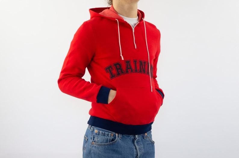 K&ouml;nnte beinhalten: Roter Kapuzenpullover mit dunkelblauem Saum und dem Schriftzug "TRAIN" in dunkelblauer Schrift auf der Vorderseite.