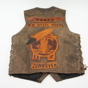 May include: Brown leather vest with a motorcycle club emblem. The vest features the text "NANCY M.C. STEEL HORSE ZONHOVEN" in a distressed style. The vest has side laces and a black lining.