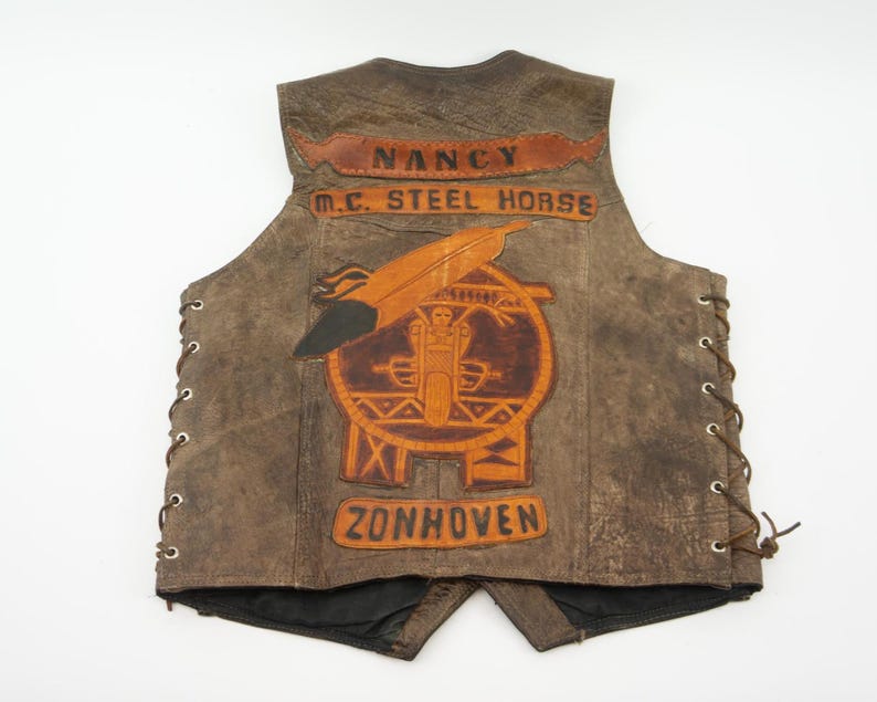 May include: Brown leather vest with a motorcycle club emblem. The vest features the text "NANCY M.C. STEEL HORSE ZONHOVEN" in a distressed style. The vest has side laces and a black lining.