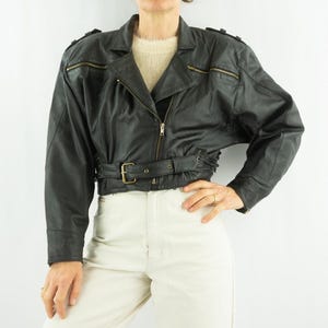 Op de afbeelding: Zwarte leren jas met een ceintuur en goudkleurige ritsen. De jas heeft een klassieke bikerstijl met schouderstukken en een reverskraag. Het model draagt de jas met een crèmekleurige broek.