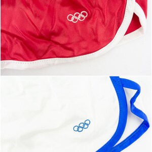 May include: Two pairs of athletic shorts with white and blue trim. One pair is red with a white Olympic rings logo. The other pair is white with a blue Olympic rings logo.